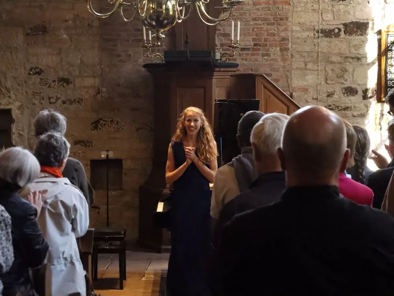 Alexandra Tchernakova smiling with her hands together as the audience applauds, in a hall with stone and brick walls and a brass chandelier