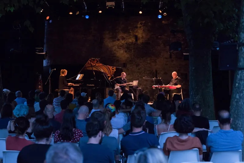 Alexandra Tchernakova at a grand piano on an outdoor stage at night with an electric guitarist and a keyboard player, the audience seated under the trees