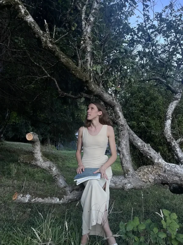 Alexandra Tchernakova in a long cream dress with a score, leaning on an old tree in a garden