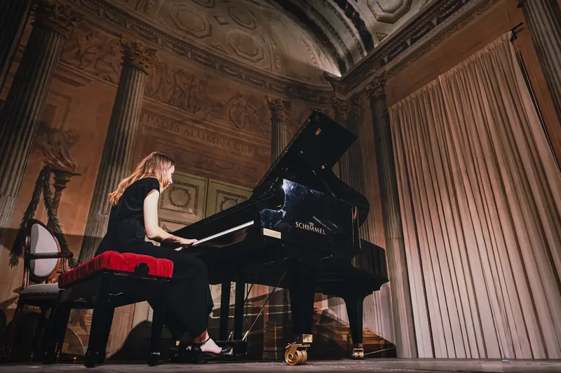 Alexandra Tchernakova playing a Schimmel grand piano in an ornate hall with columns, frescoed ceiling and draped curtains