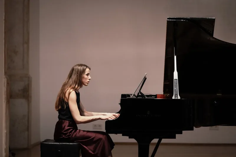Alexandra Tchernakova playing a Steinway grand piano in a bright hall with white walls and windows