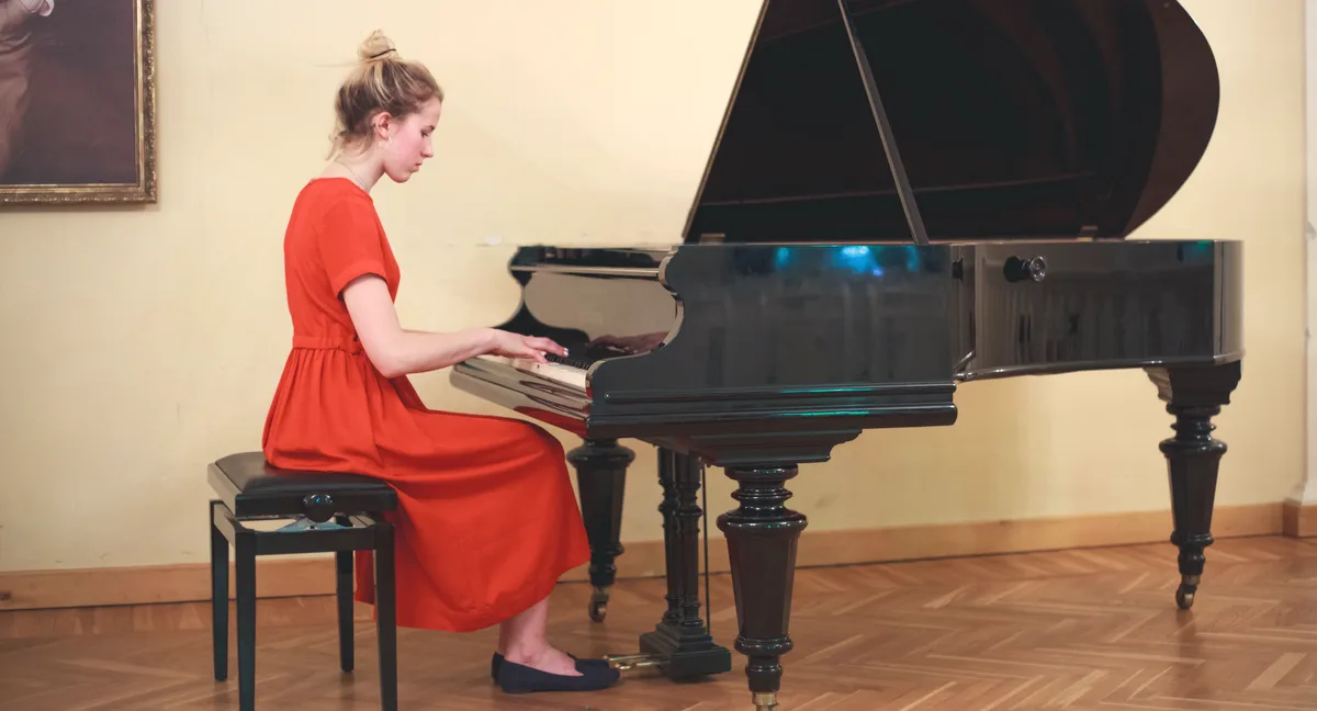 Alexandra Tchernakova in a red dress playing a black grand piano in a room with a parquet floor